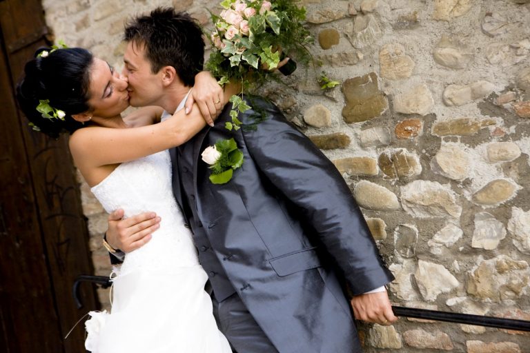 Ein Hochzeitspaar umarmt sich und küsst sich vor einer Steinmauer.