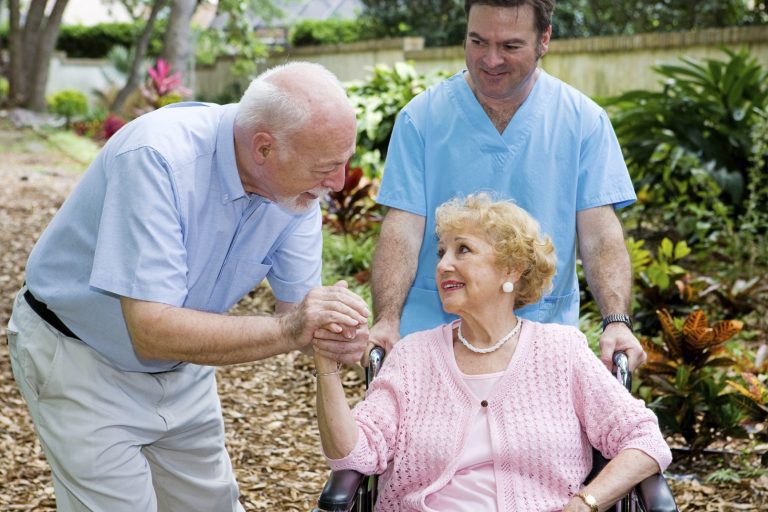 Älteres Paar in einem Garten, der Mann hält die Hand der sitzenden Frau, Betreuer im Hintergrund.