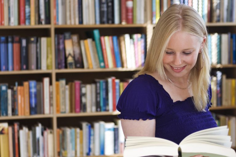 Eine lächelnde Frau blättert durch ein Buch vor einem Bücherregal.