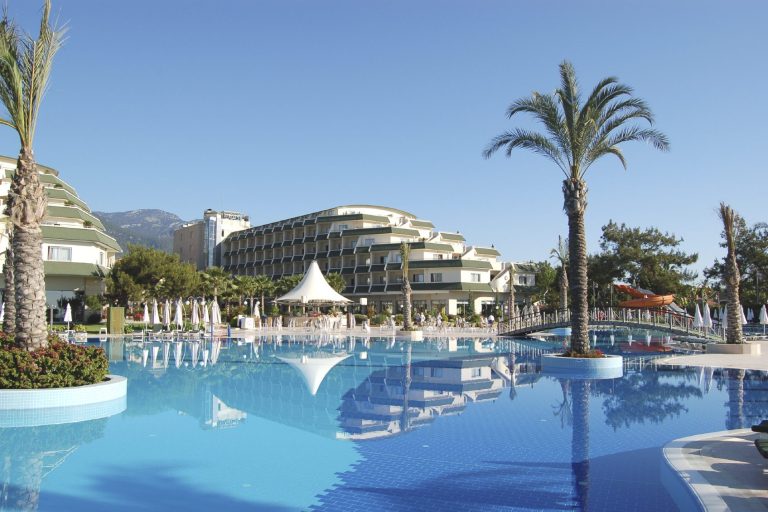 Luxuriöser Pool mit Palmen und Hotelgebäude im Hintergrund, strahlender blauer Himmel.