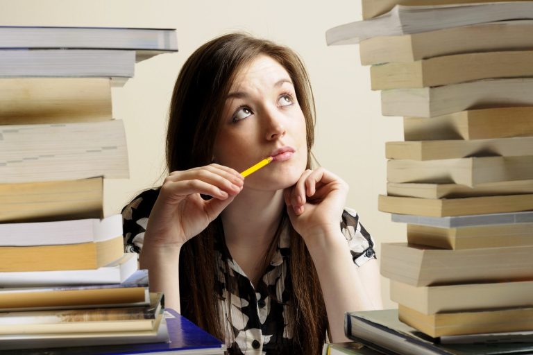 Nachdenkliche Frau mit einem Stift, umgeben von hohen Stapeln Bücher.