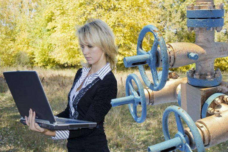 Eine Frau mit Laptop steht neben blauen Rohren und Ventilen im Freien.