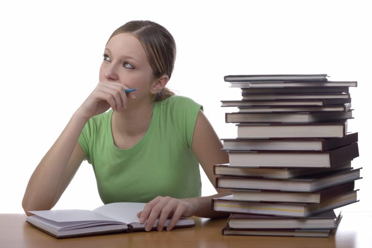 Nachdenkliche Person in grüner Bluse sitzt neben einem hohen Stapel Bücher.