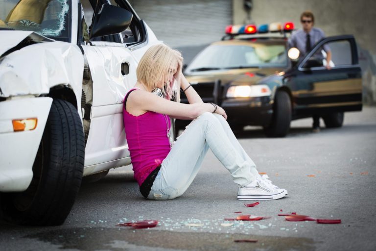 Junge Frau sitzt verzweifelt neben einem beschädigten Auto, Polizeiauto im Hintergrund.