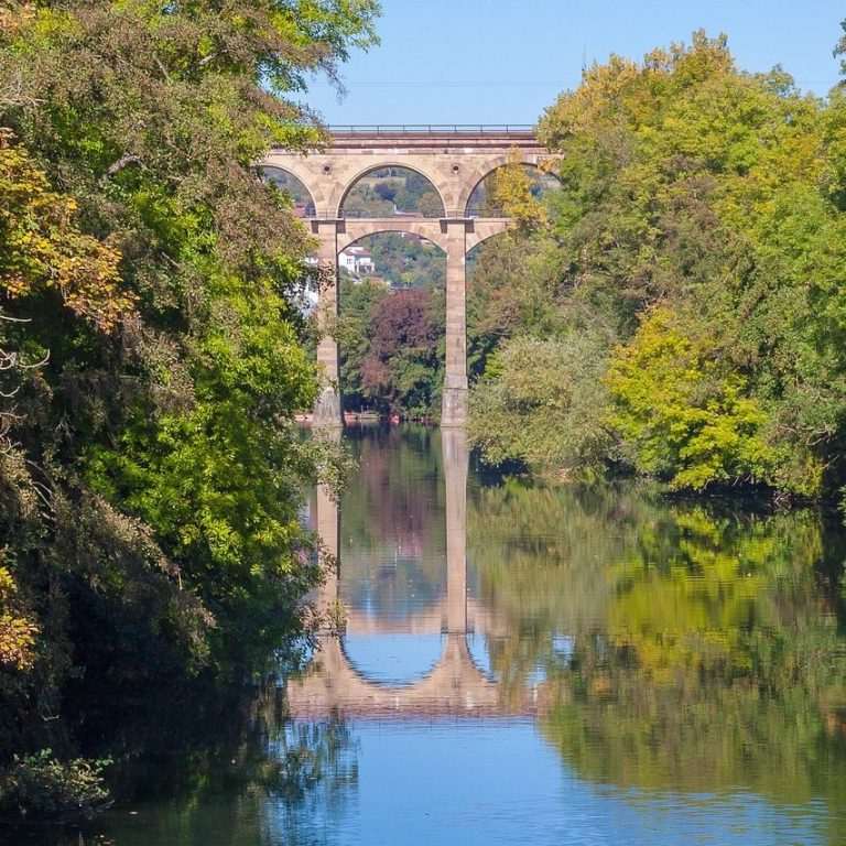 Wassertal mit grünem Ufer und reflektierendem Viadukt im Hintergrund.